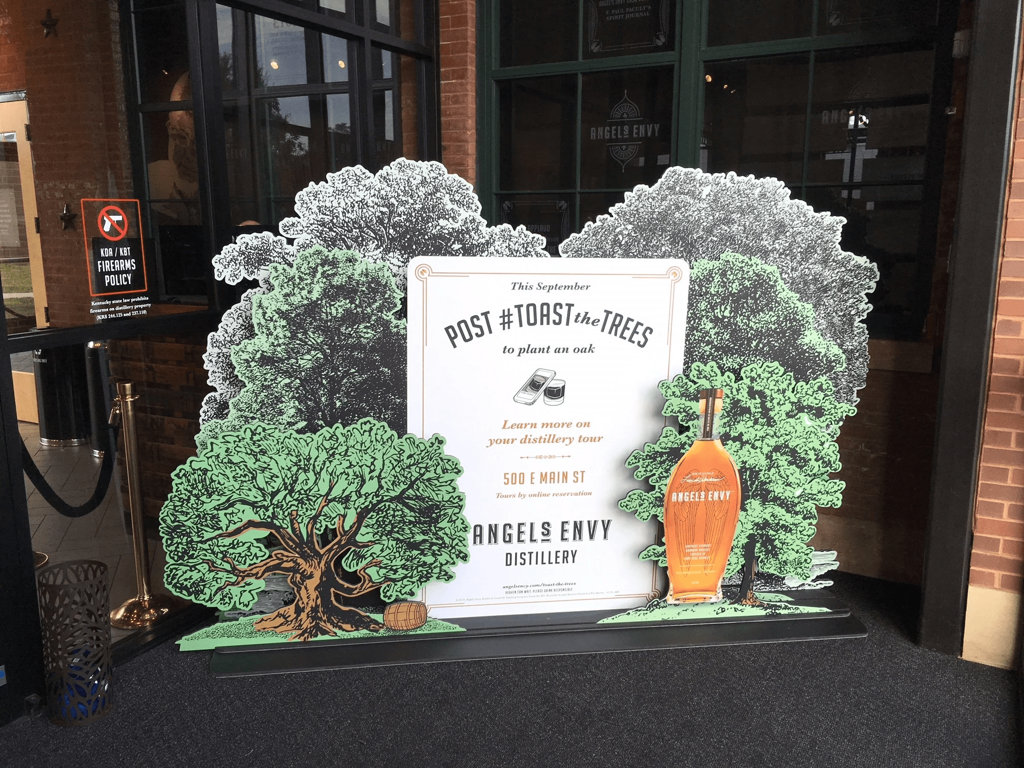 Promotional outdoor display for Angels Envy featuring a signboard with tree illustrations and a large replica of an Angels Envy bottle, next to a 'No Firearms' sign and a stanchion outside a building.