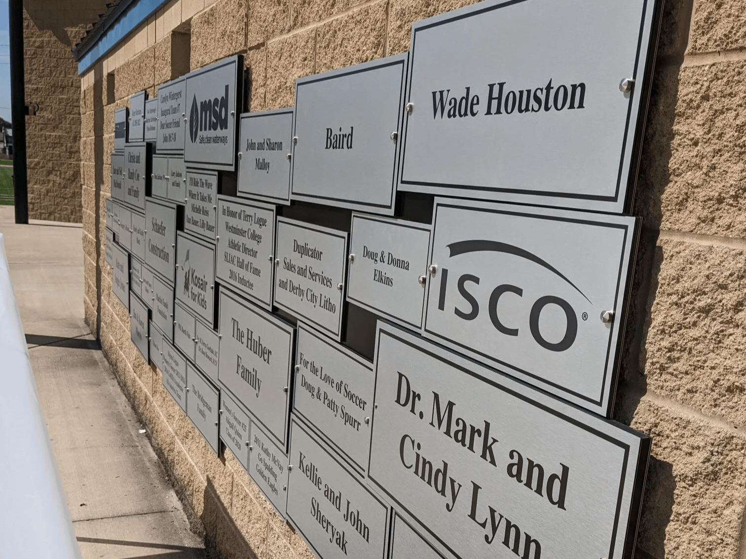 A sunny wall adorned with rows of metallic commemorative plaques bearing names and logos, suggesting recognition of donors or honored individuals.