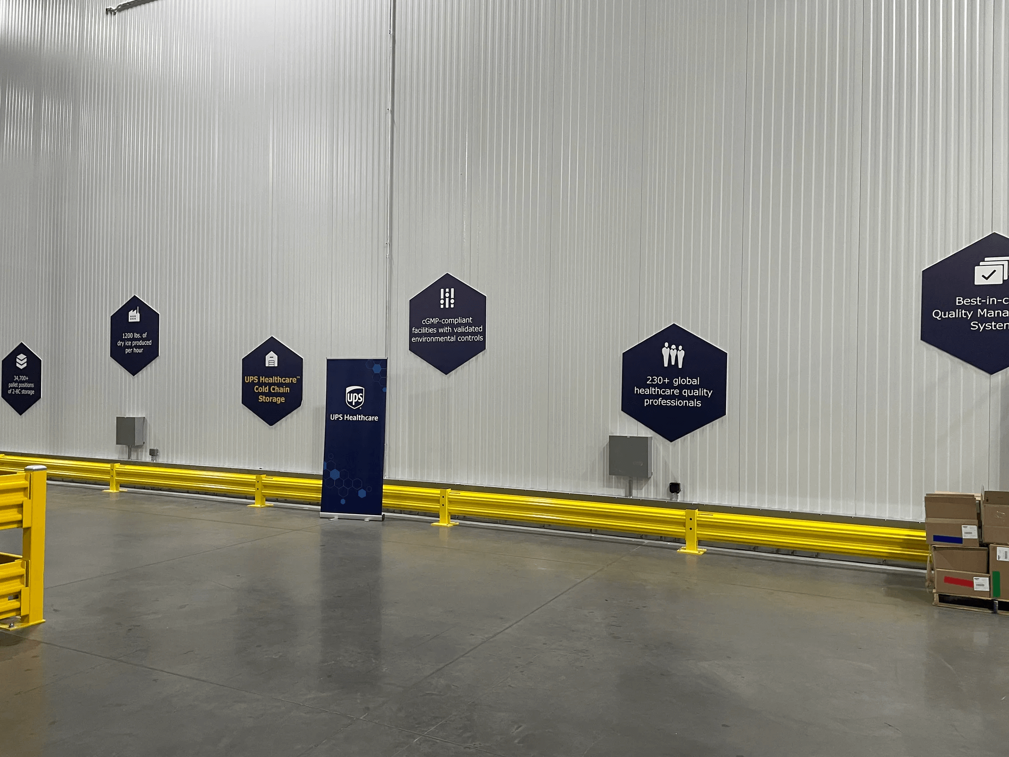 Interior of a warehouse with blue hexagon-shaped signs detailing logistics operations for healthcare on a corrugated metal wall, with a yellow safety railing on the left and a 