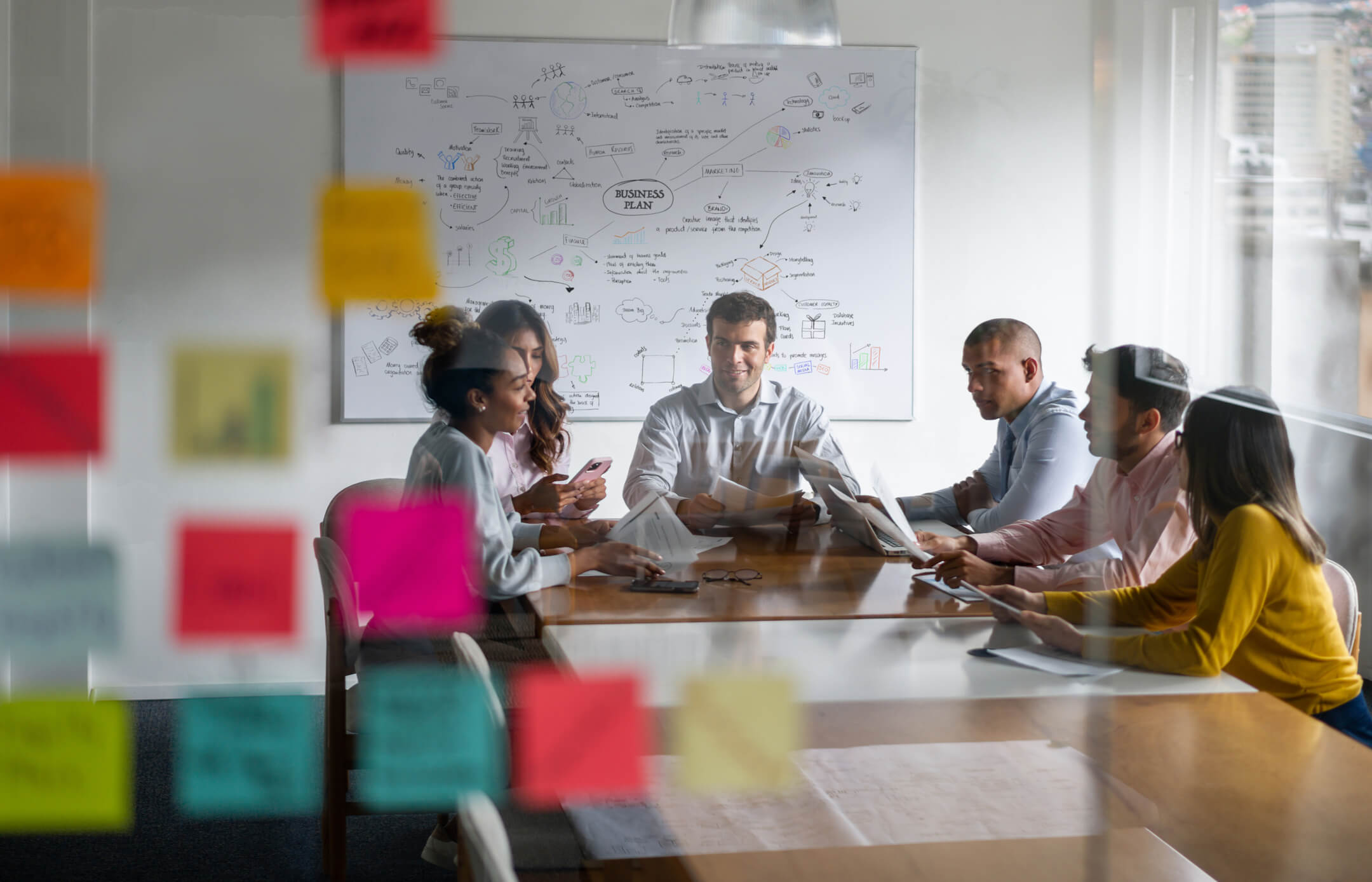 A professional team of six in a well-lit meeting room with a whiteboard full of business plans and colorful sticky notes on the window.